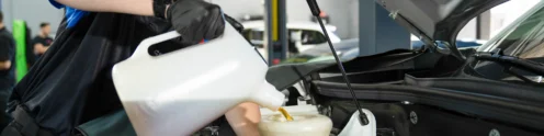 A person wearing safety glasses and gloves pours motor oil into a car engine using a funnel in an automotive workshop during a 17-Point Oil Change.