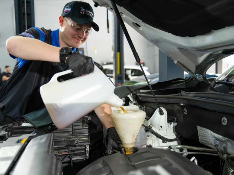 A person wearing safety glasses and gloves pours motor oil into a car engine using a funnel in an automotive workshop during a 17-Point Oil Change.