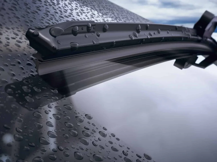 Close-up of windshield wiper blades clearing water droplets from a car windshield, with a cloudy sky reflected in the glass.