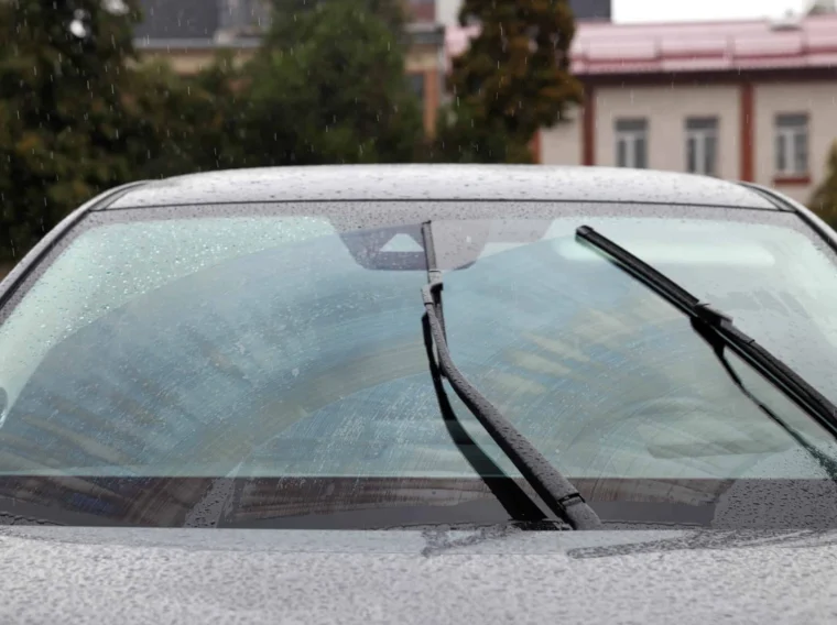 Close-up of a car windshield with windshield wiper blades in motion, clearing raindrops during a rainy day; buildings are visible in the blurred background.