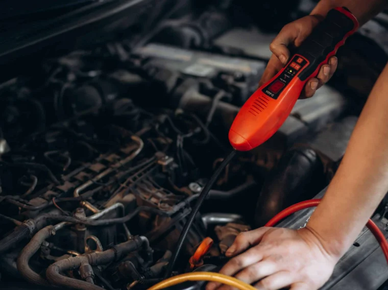 Person using a red diagnostic tool under a car hood, inspecting engine components with cables connected—part of comprehensive A/C services to ensure your vehicle’s air conditioning system runs smoothly.