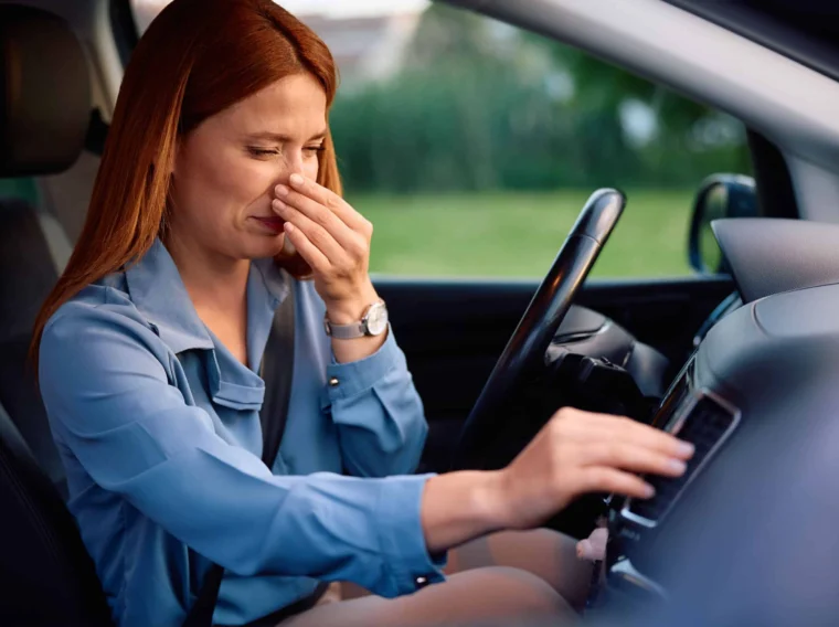 A woman sitting in a car pinches her nose with one hand and adjusts the air conditioning controls with the other, possibly reacting to an unpleasant smell after a recent clutch replacement.
