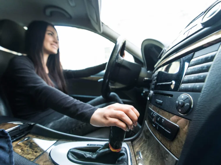 A woman sits in the driver's seat of a car, holding the steering wheel with one hand and shifting the gear lever with the other after a recent clutch replacement.