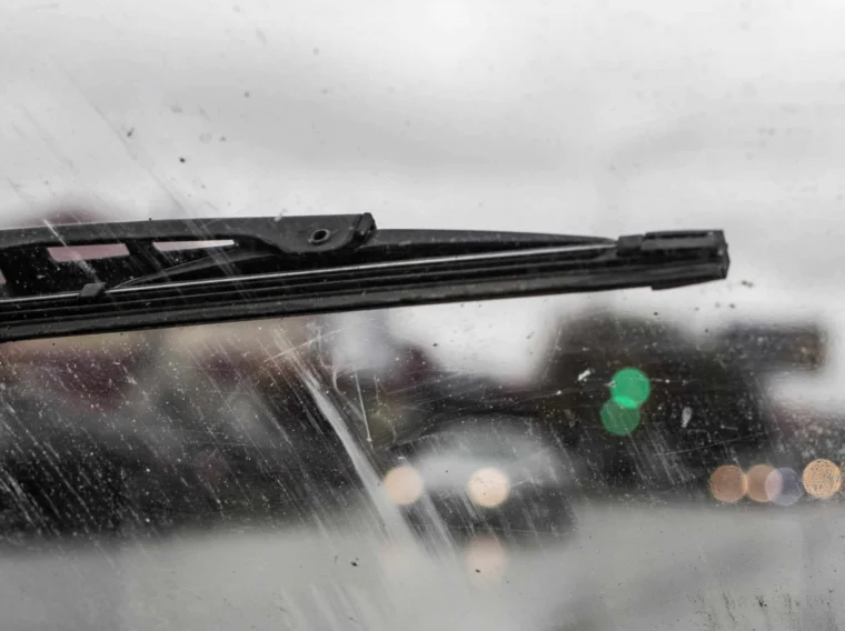 A close-up of windshield wiper blades on a dirty car window, with blurred cars and streetlights in the background on a cloudy day.