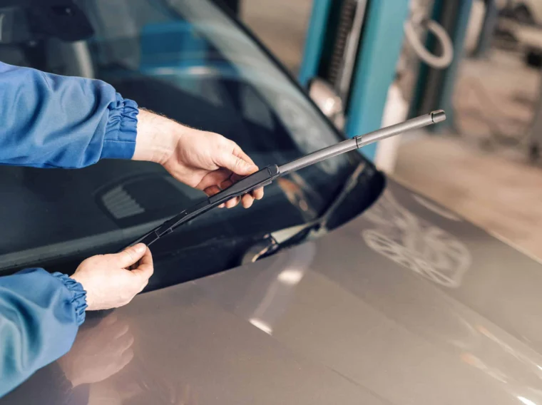 A person replaces windshield wiper blades on a car in a garage or workshop setting.