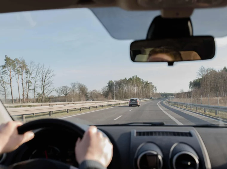 View from inside a car driving on a two-lane highway lined with trees, another car ahead in the distance. The driver's hands are on the wheel—reminding the importance of regular brake maintenance for safe journeys.