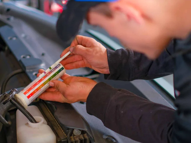 A person wearing a black cap uses a coolant tester to check the coolant in a car engine bay, demonstrating careful radiator maintenance.