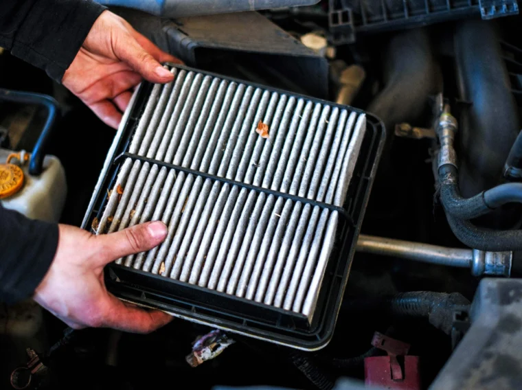 A person’s hands are removing or installing a rectangular car air filter with visible dirt and debris from an engine compartment, showcasing essential filter services.