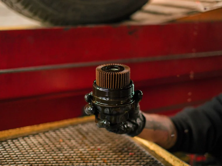 A person wearing a black glove holds a used, oil-covered engine oil filter in a garage setting, highlighting the importance of regular Filter Services.