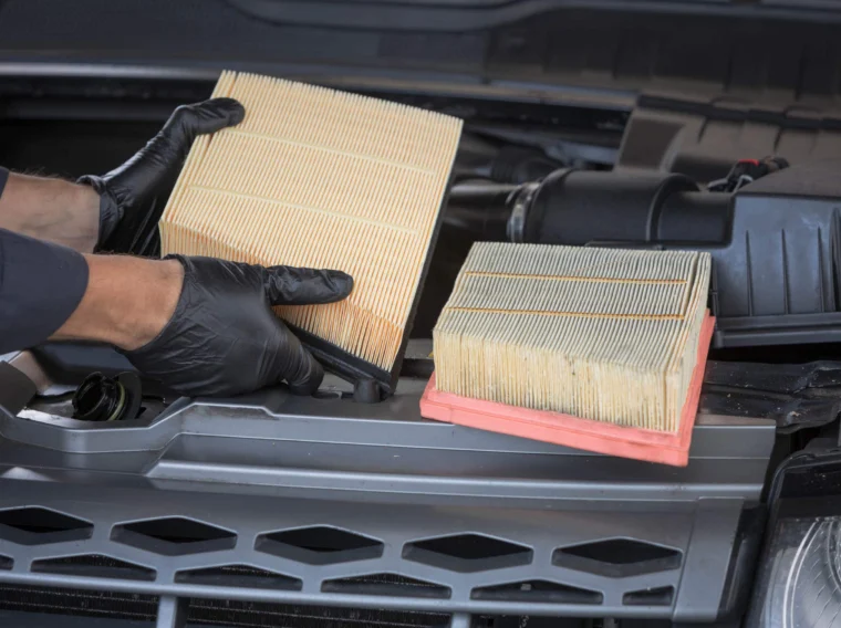 A person wearing black gloves holds a new engine air filter next to an old, dirty filter under the hood of a car, highlighting the importance of regular Filter Services.