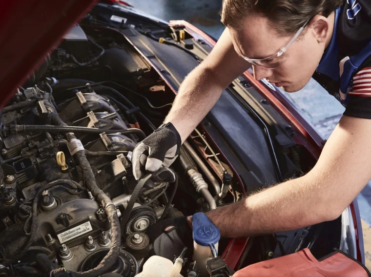 A mechanic wearing gloves and safety glasses works on the engine of a car with the hood open.