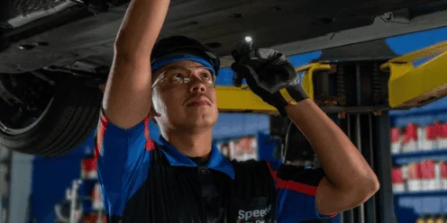 A mechanic wearing safety glasses and gloves uses a flashlight to inspect the underside of a raised vehicle in a garage.