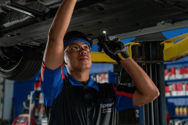 A mechanic wearing safety glasses and gloves uses a flashlight to inspect the underside of a raised vehicle in a garage.