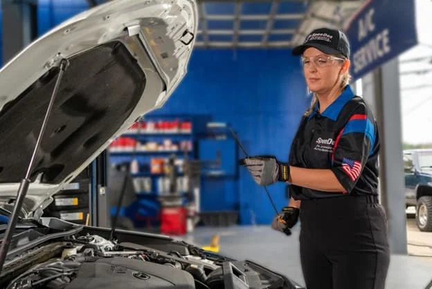 An auto technician wearing safety gear holds a dipstick while inspecting a car engine in a service garage.
