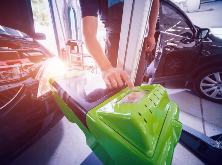 A mechanic operates a green diagnostic machine next to a black car in an auto repair shop, with sunlight and expert lighting services brightening the workspace through the window.