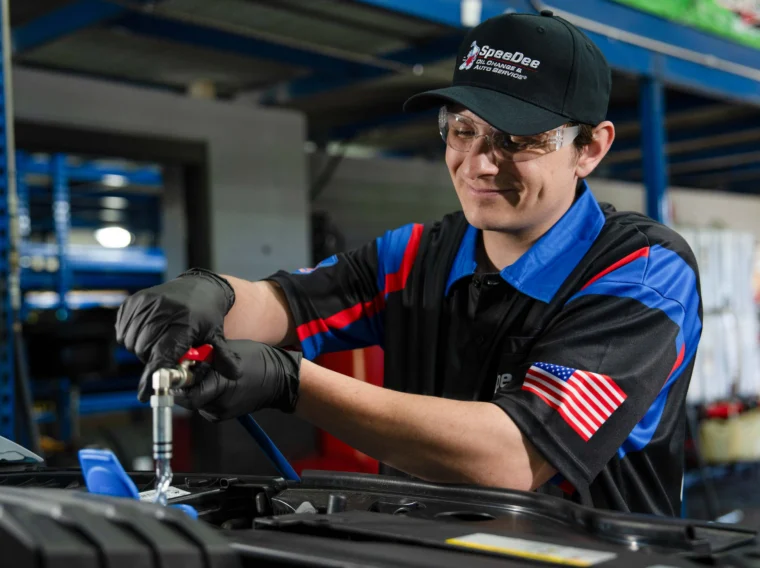 A mechanic in uniform and safety glasses performs leak diagnostics under a car hood in a garage, with shelves and equipment visible in the background.