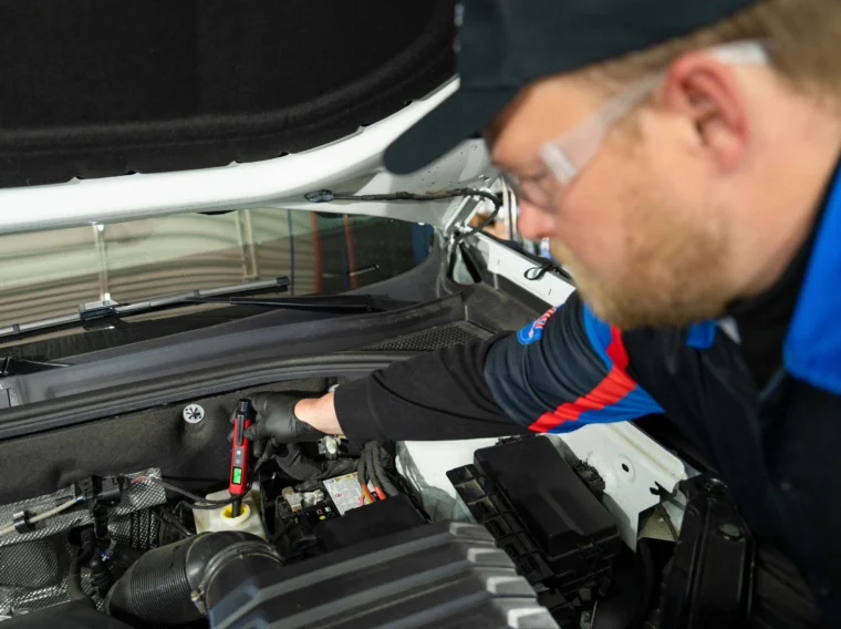 A mechanic wearing gloves and safety glasses checks under the hood of a vehicle, using a diagnostic tool for leak diagnostics near the engine components.