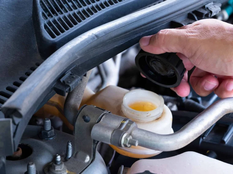 A hand is opening the brake fluid reservoir cap in a car engine bay, showing the fluid level inside the container.