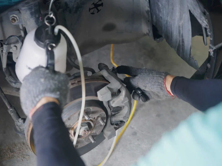 Person wearing gloves uses a tool to bleed the brakes on a car, with a tube connected to a brake bleeder bottle attached to the brake caliper.