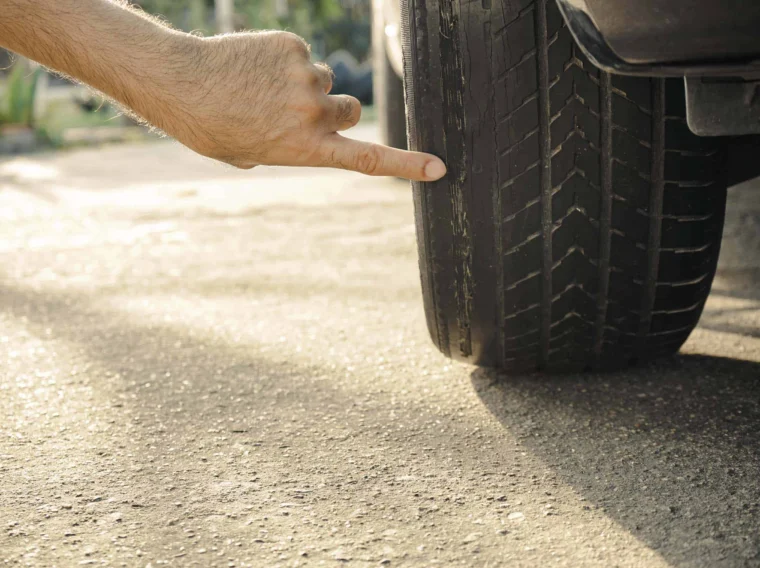 A hand points to the worn tread on a car tire, highlighting significant tire wear and the urgent need for tire replacement.
