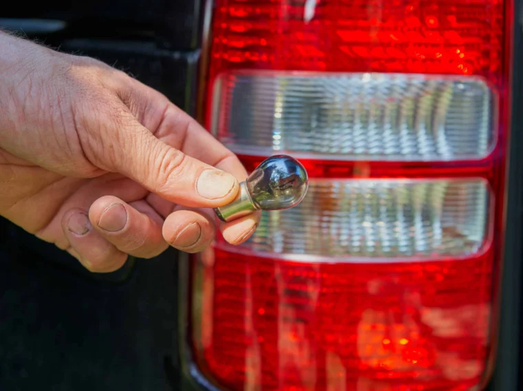 A hand holds a car tail light bulb in front of a vehicle's rear tail light assembly, demonstrating expert Lighting Services.
