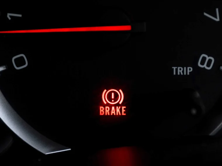 Close-up of a car dashboard showing a red brake warning indicator light illuminated next to the word "BRAKE," with part of the speedometer and trip meter visible.