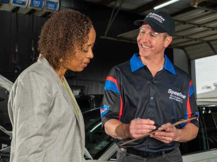 A mechanic in uniform holds a clipboard and smiles while discussing a check engine light issue with a woman in a blazer inside an auto repair shop.