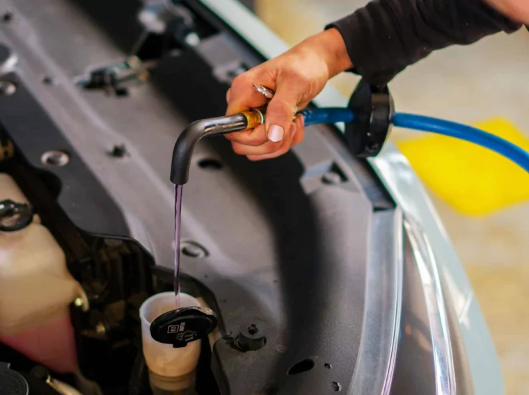 A person is performing a fluid exchange, filling a car's coolant reservoir with liquid using a hose under the hood of the vehicle.