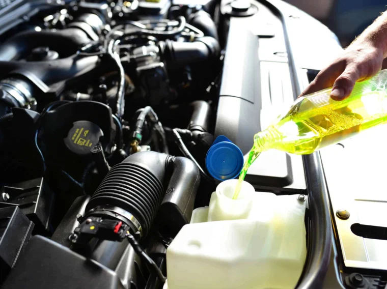 A person pours yellow-green fluid engine coolant, into a car's engine compartment reservoir as part of a routine fluid exchange.