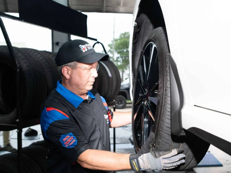 A mechanic wearing gloves and uniform is installing a tire on a white vehicle in an auto shop, with a rack of tires in the background—demonstrating expert care and readiness for State Inspection Services.