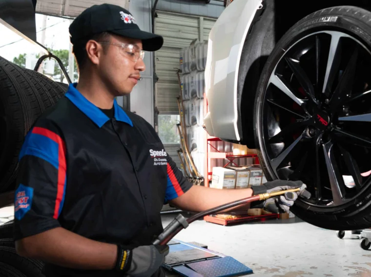 An auto mechanic in uniform provides Tire Services by inflating a car tire with an air hose in a garage. The car sits on a hydraulic lift, surrounded by various equipment in the background.