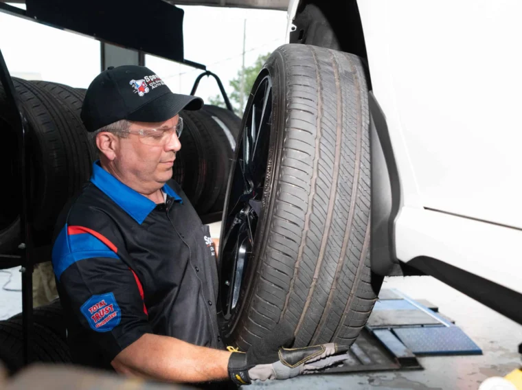 A mechanic in uniform and safety glasses lifts a car tire onto a raised vehicle, expertly handling Tire Replacement in an auto repair shop.
