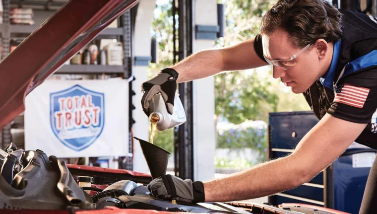 A mechanic wearing gloves and safety glasses pours engine oil into a car’s engine bay using a funnel in an auto shop.