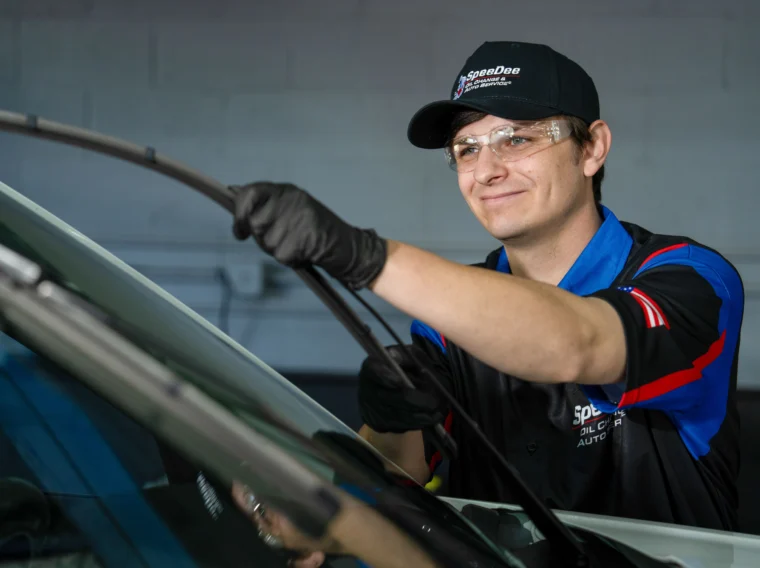 A mechanic wearing gloves, safety glasses, and a cap is replacing a windshield wiper on a car during routine tune up services in an auto repair shop.