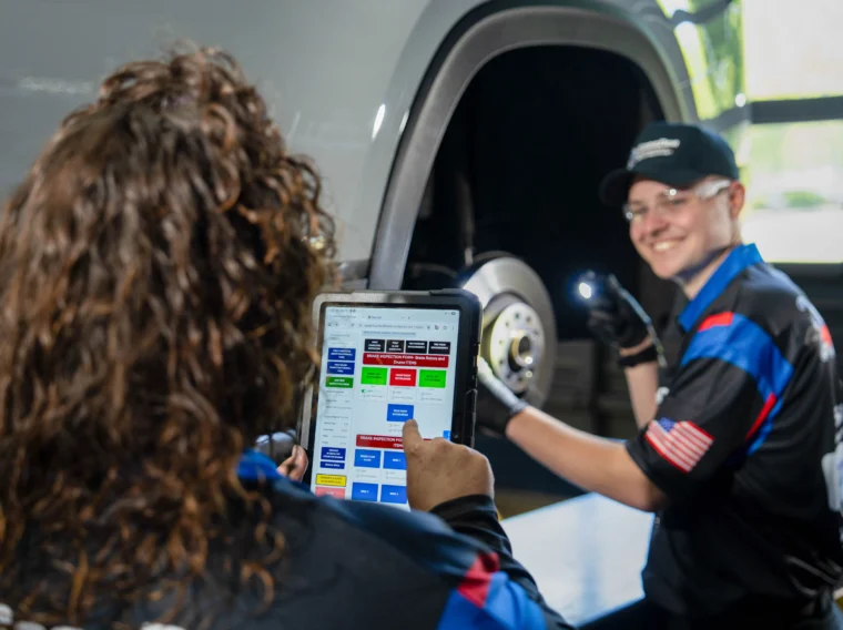 A person uses a tablet with a diagnostic interface while another inspects a vehicle's brake system in a garage, ensuring all maintenance—from brakes to Alignment Services—is expertly managed.