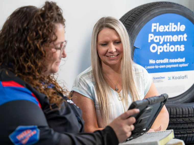 Two women sit near tires; one shows the other something on a tablet, possibly discussing Alignment Services. A sign in the background reads “Flexible Payment Options, No credit needed.”.