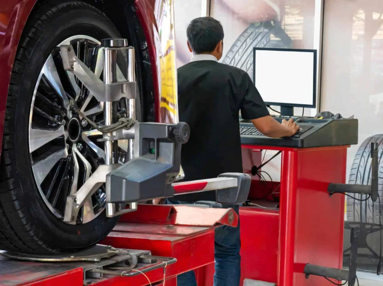 A technician stands at a computer workstation while a car, positioned on a lift with a wheel alignment tool attached to its front tire, receives expert Alignment Services.