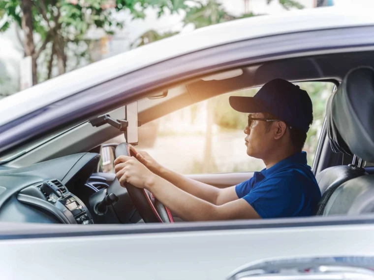 A man wearing a blue shirt and cap sits in the driver's seat of a parked car, holding the steering wheel and looking forward—perhaps awaiting professional Alignment Services.