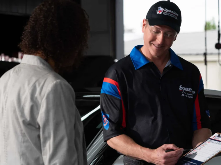 A mechanic in a Speed Auto Repair shirt shows a clipboard to a customer standing beside a car in a garage.