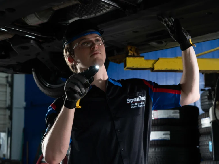 An auto mechanic inspects the underside of a vehicle with a flashlight while wearing safety glasses and gloves in a garage.
