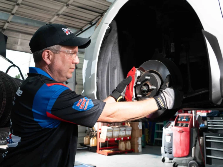 Mechanic wearing safety glasses and gloves installs a brake disc on the front wheel of a raised vehicle in an auto repair shop.