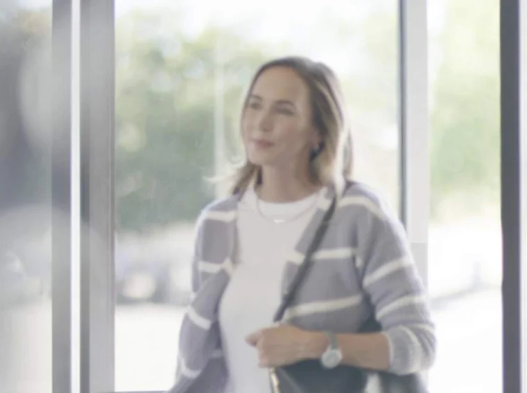 A woman with shoulder-length hair, wearing a striped cardigan and carrying a bag, stands indoors near large windows with sunlight streaming in.