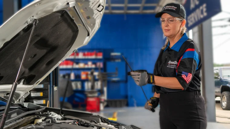 A mechanic in uniform stands next to an open car hood, holding a dipstick to check the engine oil inside an automotive service garage.