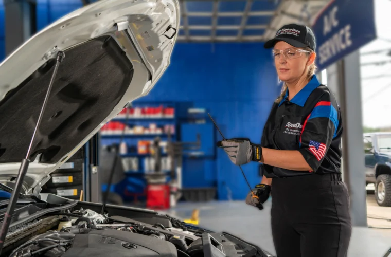 A mechanic in uniform stands next to an open car hood, holding a dipstick to check the engine oil inside an automotive service garage.