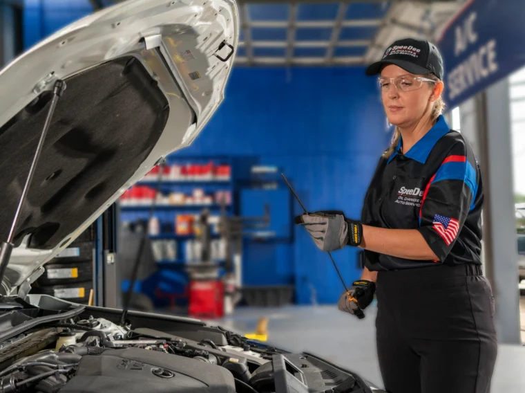 A mechanic in uniform stands next to an open car hood, holding a dipstick to check the engine oil inside an automotive service garage.