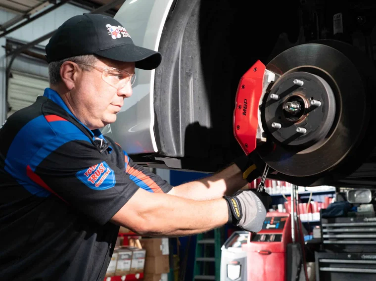 A mechanic wearing safety glasses performs Manufacturer’s Maintenance on a car’s brake system in an auto repair shop.