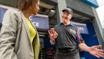 A woman in a gray blazer talks to a smiling man in a mechanic uniform and cap who is making an "OK" gesture inside an automotive service garage.