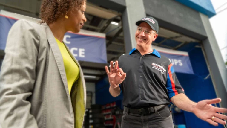 A woman in a gray blazer talks to a smiling man in a mechanic uniform and cap who is making an "OK" gesture inside an automotive service garage.