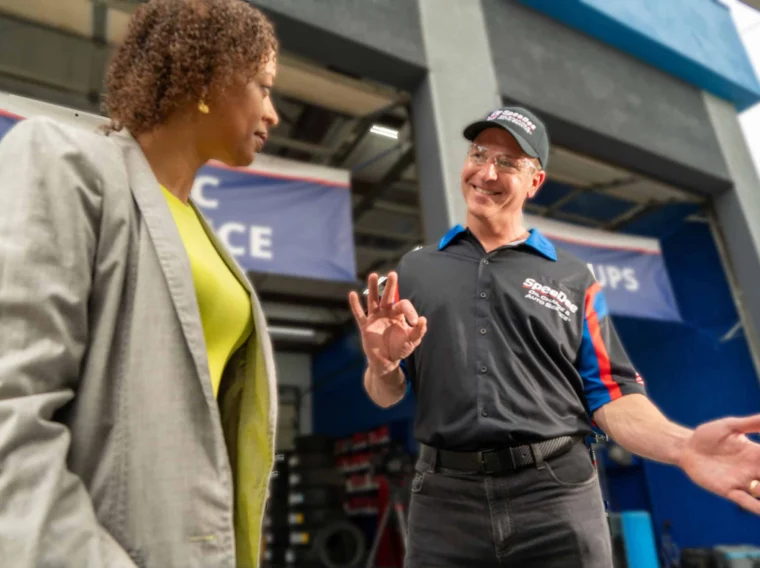 A woman in a gray blazer talks to a smiling man in a mechanic uniform and cap who is making an "OK" gesture inside an automotive service garage.