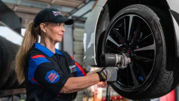 A mechanic wearing safety glasses and gloves uses a power tool to work on a car tire in an auto repair shop.